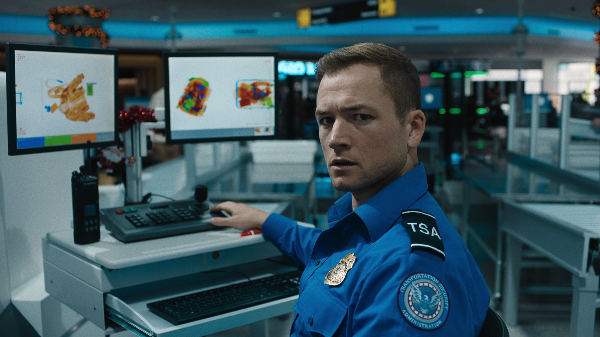 Taron Egerton as a TSA agent sitting in front of a baggage scanning screen in Carry-On